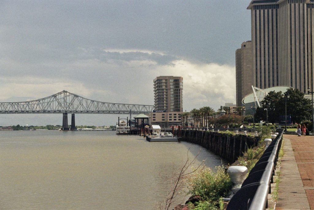 View of the Mississippi River from New Orleans on an overcast day. Green foliage line the port as people walk along the shore. The the Crescent City Connector is featured.